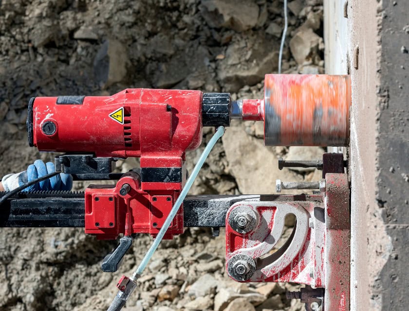 Ein Arbeiter bohrt mit einer Kernbohrmaschine in eine Betonwand. Kernbohrer, die in Metall verwendet werden, heißen Ringfräser. Kernbohrer für Beton und Hartgestein verwenden in der Regel Industrie-Diamantkorn.